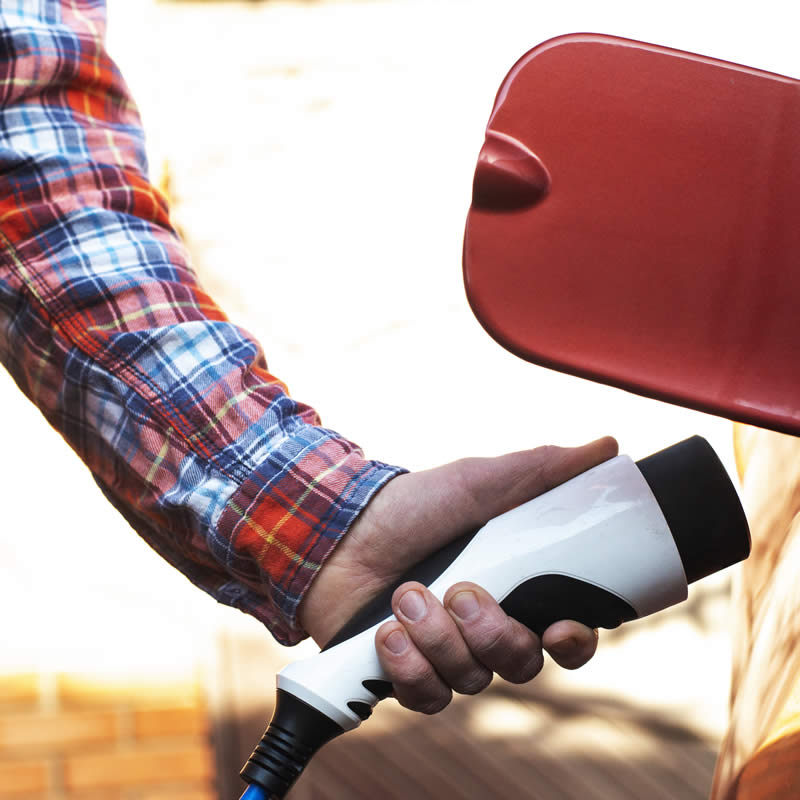 Man holding an electric vehicle charging plug near a car's charging port, illustrating home EV charger installation and maintenance.