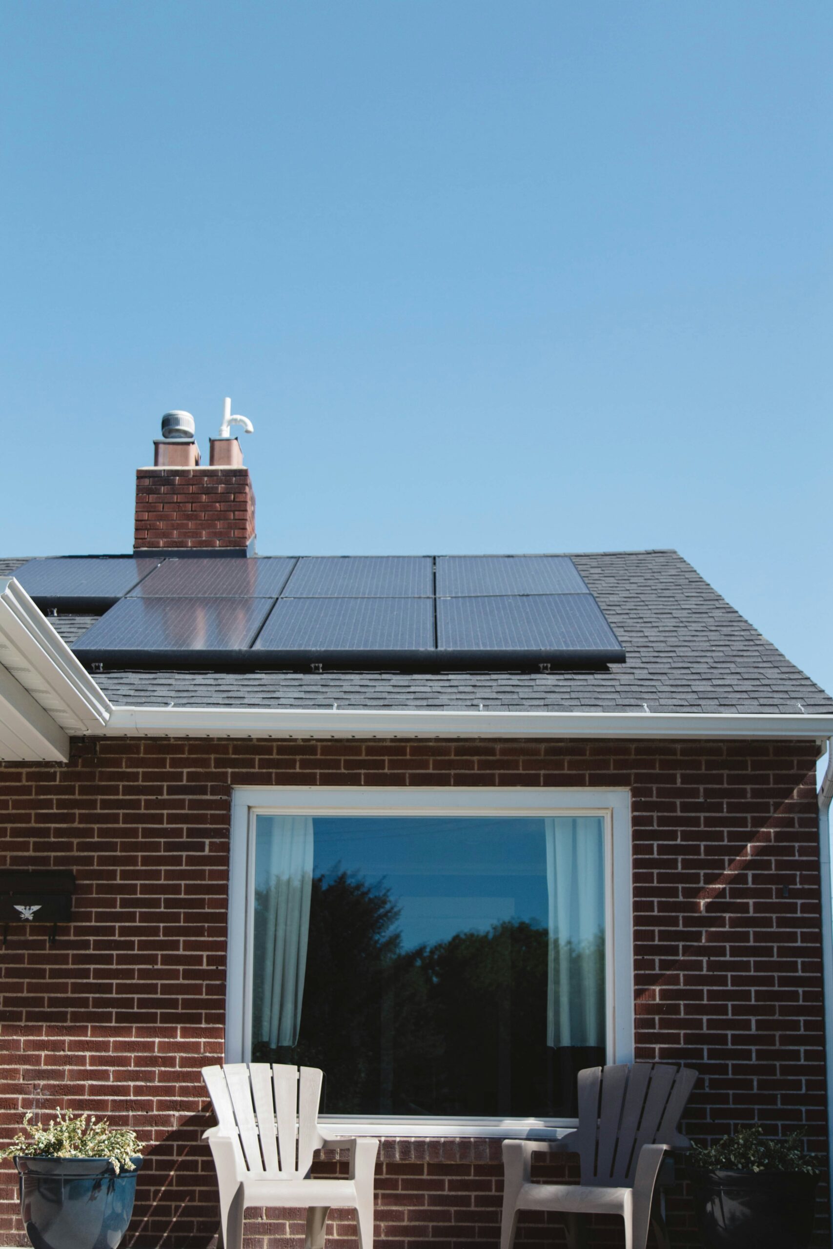 Solar panels installed on a residential roof with a brick exterior, featuring a chimney, window, and outdoor seating area, highlighting sustainable energy solutions for home EV charging.