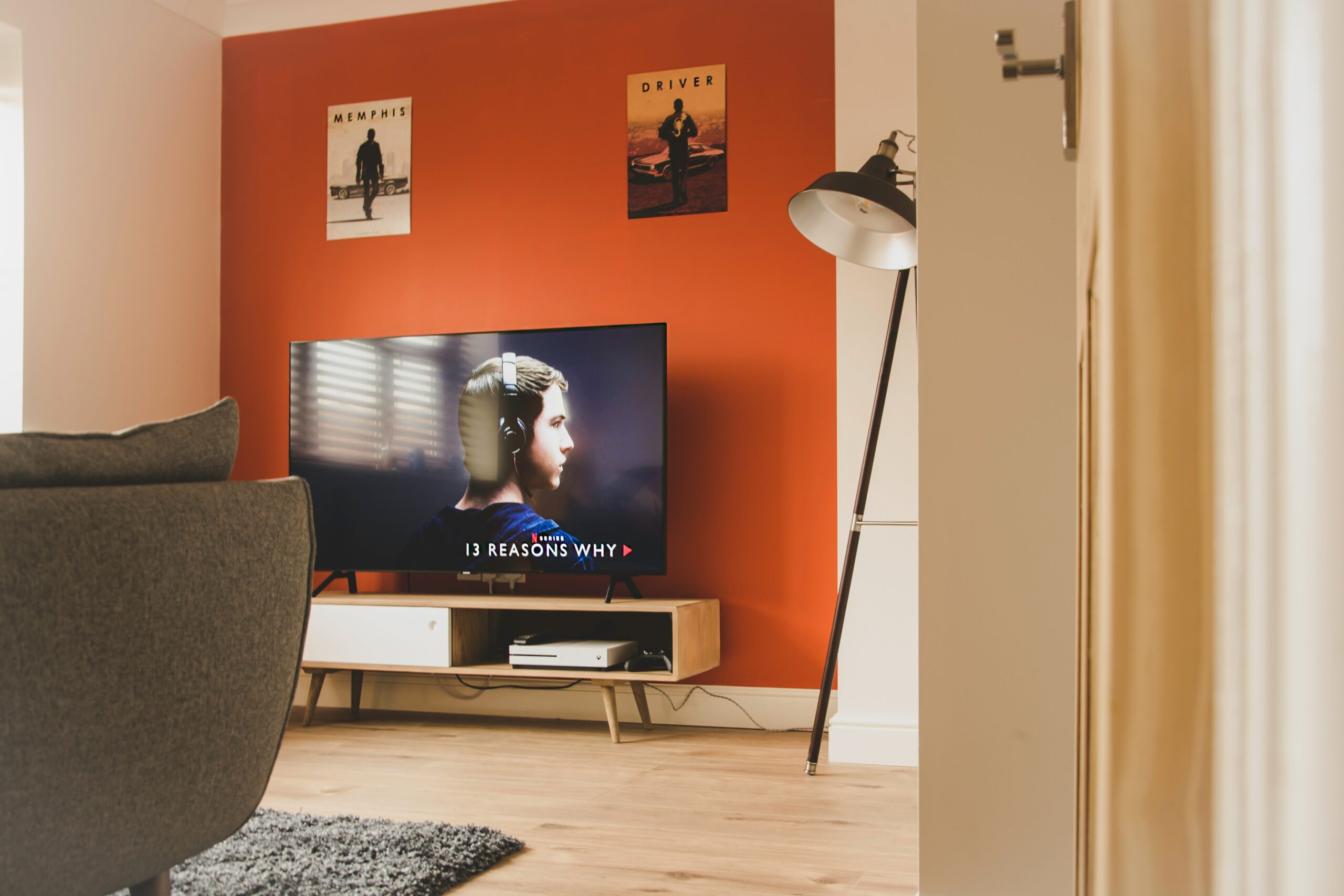 Living room featuring a television displaying "13 Reasons Why", accompanied by a grey armchair, a stylish floor lamp, and a vibrant orange wall with movie posters.