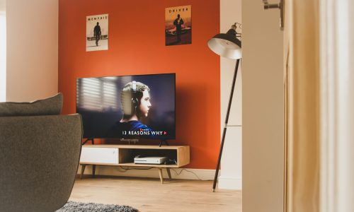 Modern living room featuring a television displaying "13 Reasons Why", stylish furniture, and a warm orange accent wall, highlighting automated lighting control for enhanced comfort.