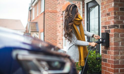 Woman in a grey sweater and yellow scarf connecting an electric vehicle to a home EV charger on a brick wall, with a car partially visible in the foreground.